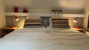 A bedroom with a bed, two bedside tables, two lamps, a shelf with decorative items, and a window.