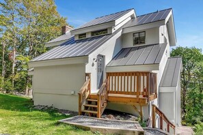 A two-story house with a sloped metal roof, a wooden deck, and a small staircase leading to the upper level.