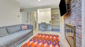 Un salon moderne avec un canapé gris, un tapis coloré, un téléviseur fixé à un mur en briques et une bibliothèque.