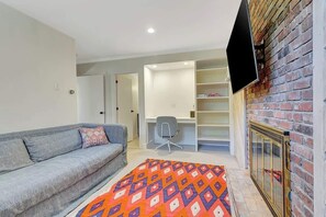 Un salon moderne avec un canapé gris, un tapis coloré, un téléviseur fixé à un mur en briques et une bibliothèque.