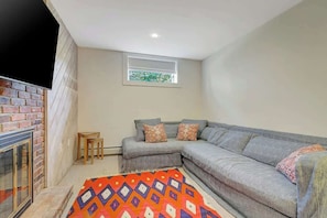 A living room with a grey sectional sofa, a small wooden table, a colorful rug, and a brick fireplace.