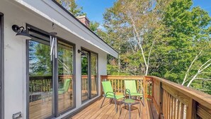 Une terrasse en bois avec des portes et des fenêtres en verre, des chaises vertes et une vue sur les arbres.
