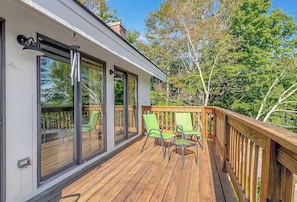 A wooden deck with glass doors and windows, green chairs, and a view of trees.