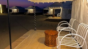 Un campamento en el desierto con tiendas de campaña, una zona de estar con sillas y una mesa, y vistas del desierto al anochecer.