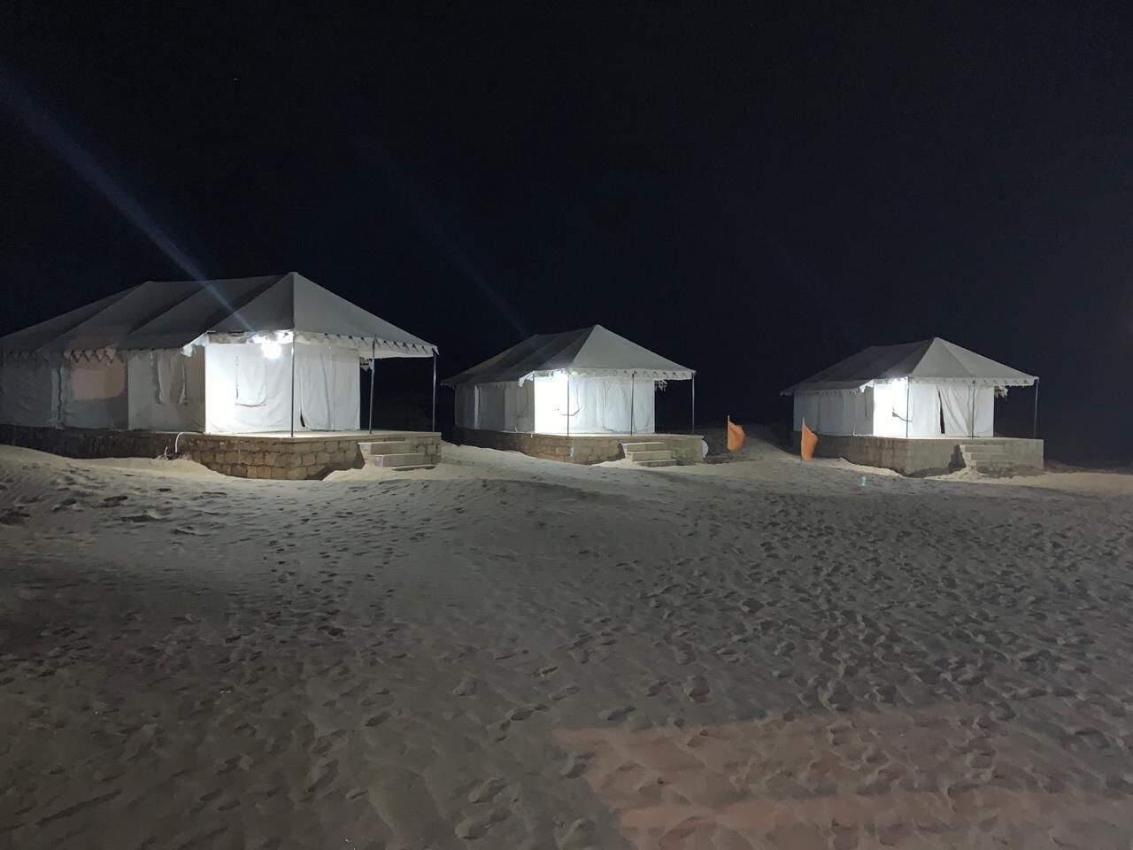 Tres tiendas blancas iluminadas sobre una superficie arenosa, de noche.