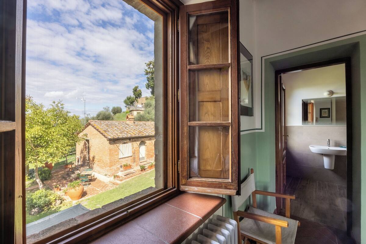 A room with a large window, a wooden bench, a chair, and a view of a garden and a building outside.