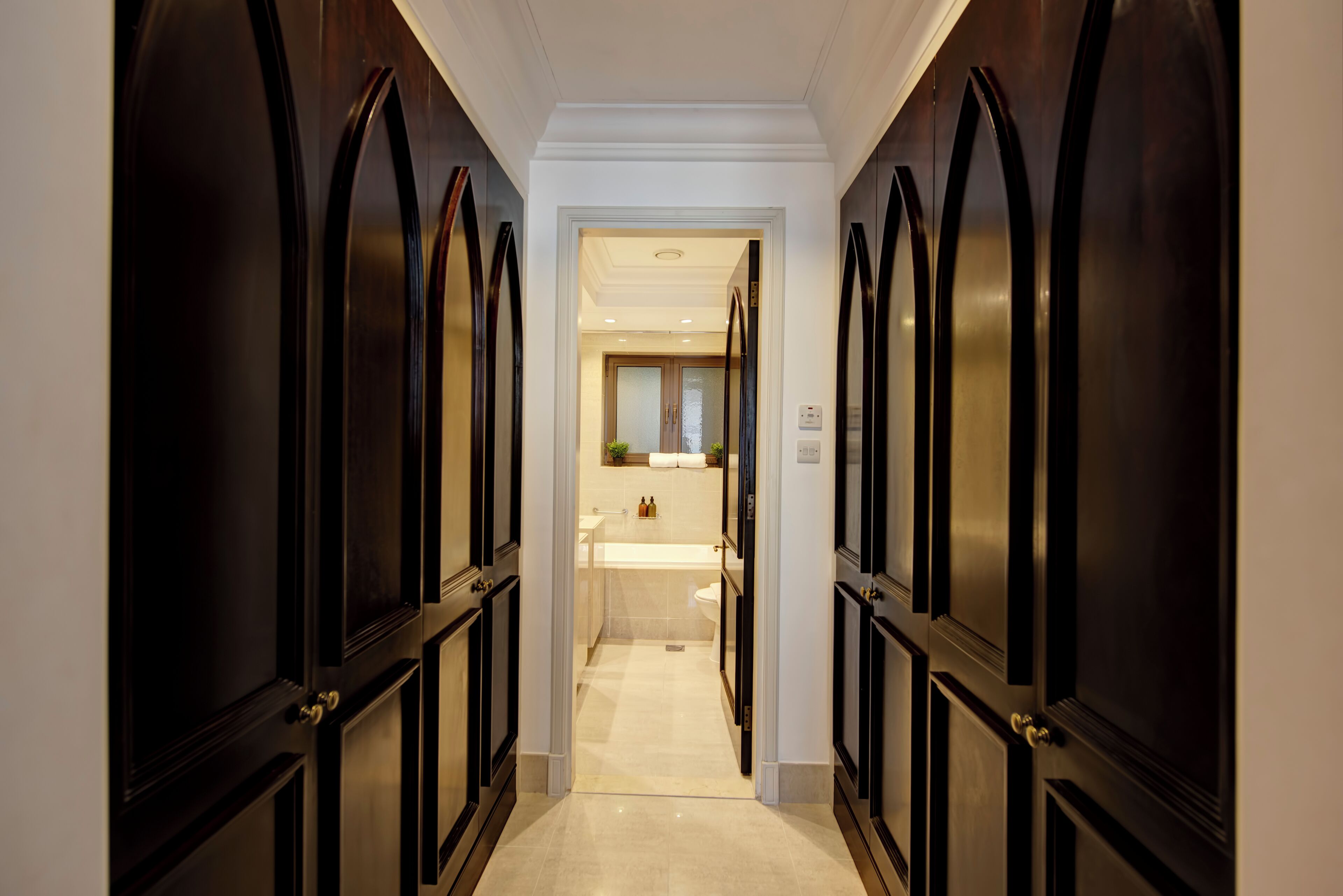 Un couloir avec des portes en bois foncé menant à une salle de bain équipée d’une baignoire et de toilettes.