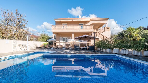 A swimming pool with a clear blue tile, a two-story building with balconies, and outdoor furniture.