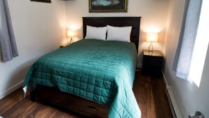 A neatly made bed with a green quilt, two bedside tables with lamps, a painting above the bed, and a small shelf with items on the wall.
