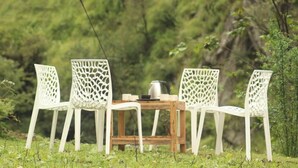 Uma mesa de madeira com quatro cadeiras brancas com desenhos recortados detalhados, colocada em uma área gramada com árvores ao fundo.