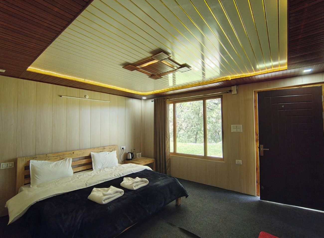 A hotel room with a wooden ceiling, a large bed with white linens, a bedside table with a lamp, and a window with a view of greenery.