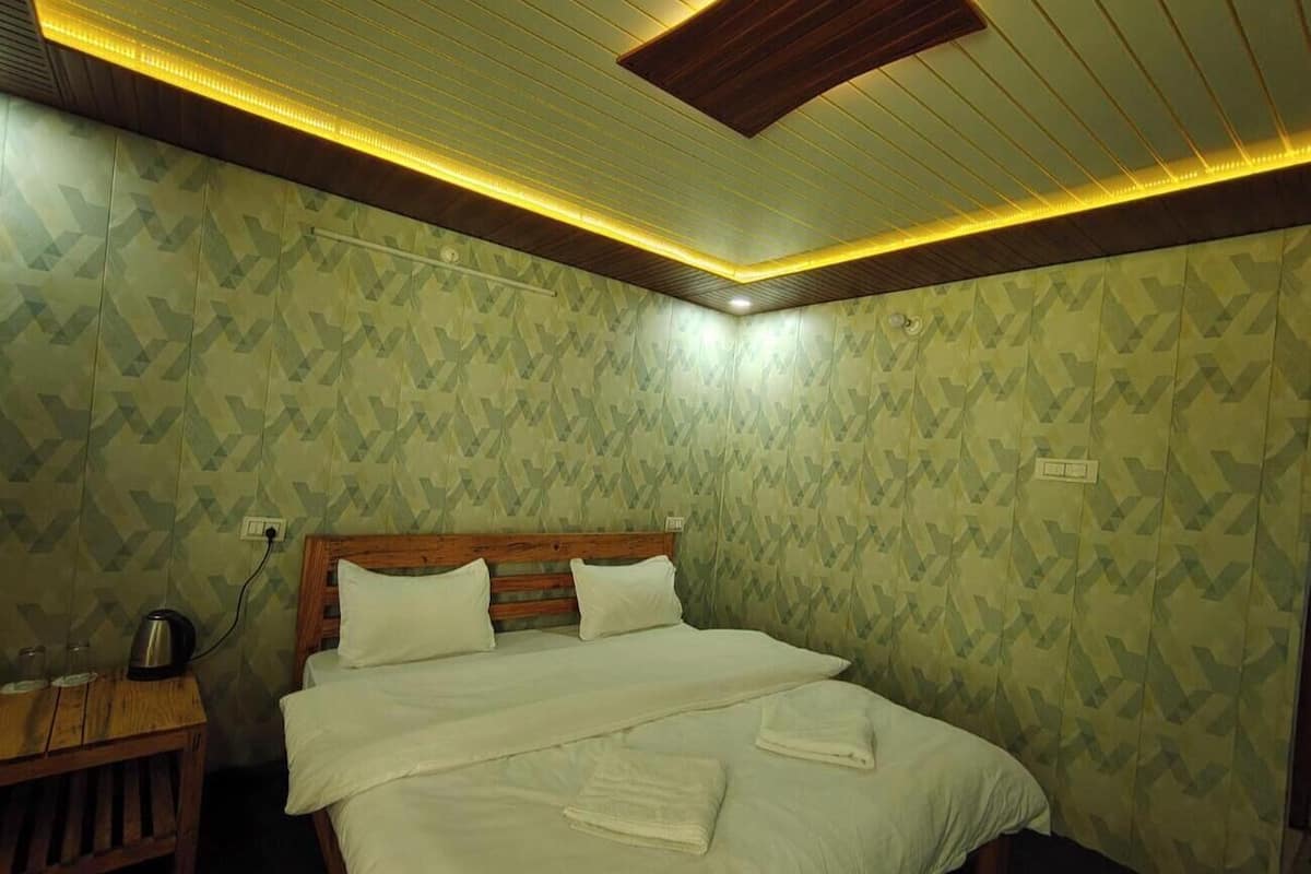 A neatly made bed with white linens and pillows, a wooden headboard, and a bedside table with a kettle. The room has patterned wallpaper and a wooden ceiling with recessed lighting.