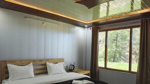 Quarto com uma cama grande, cabeceira de madeira, mesa de cabeceira com chaleira e janela com vista para árvores.