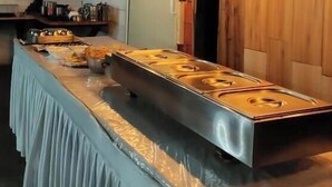 Restaurante com balcão extenso, luminárias pendentes e uma televisão exibindo um artista.