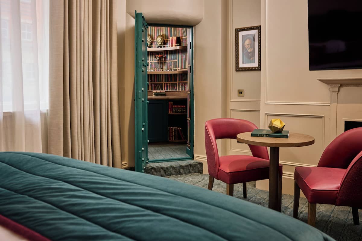 Une chambre d’hôtel avec un lit, deux fauteuils rouges, une petite table avec un livre, une bibliothèque et un téléviseur fixé au mur.