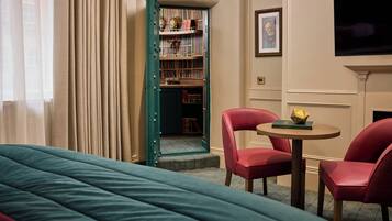 A hotel room with a bed, two red chairs, a small table with a book, a bookshelf, and a TV mounted on the wall.