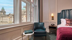 Une chambre d’hôtel avec vue sur un bâtiment historique, un fauteuil confortable, une table de chevet avec une lampe et un lit soigneusement fait.
