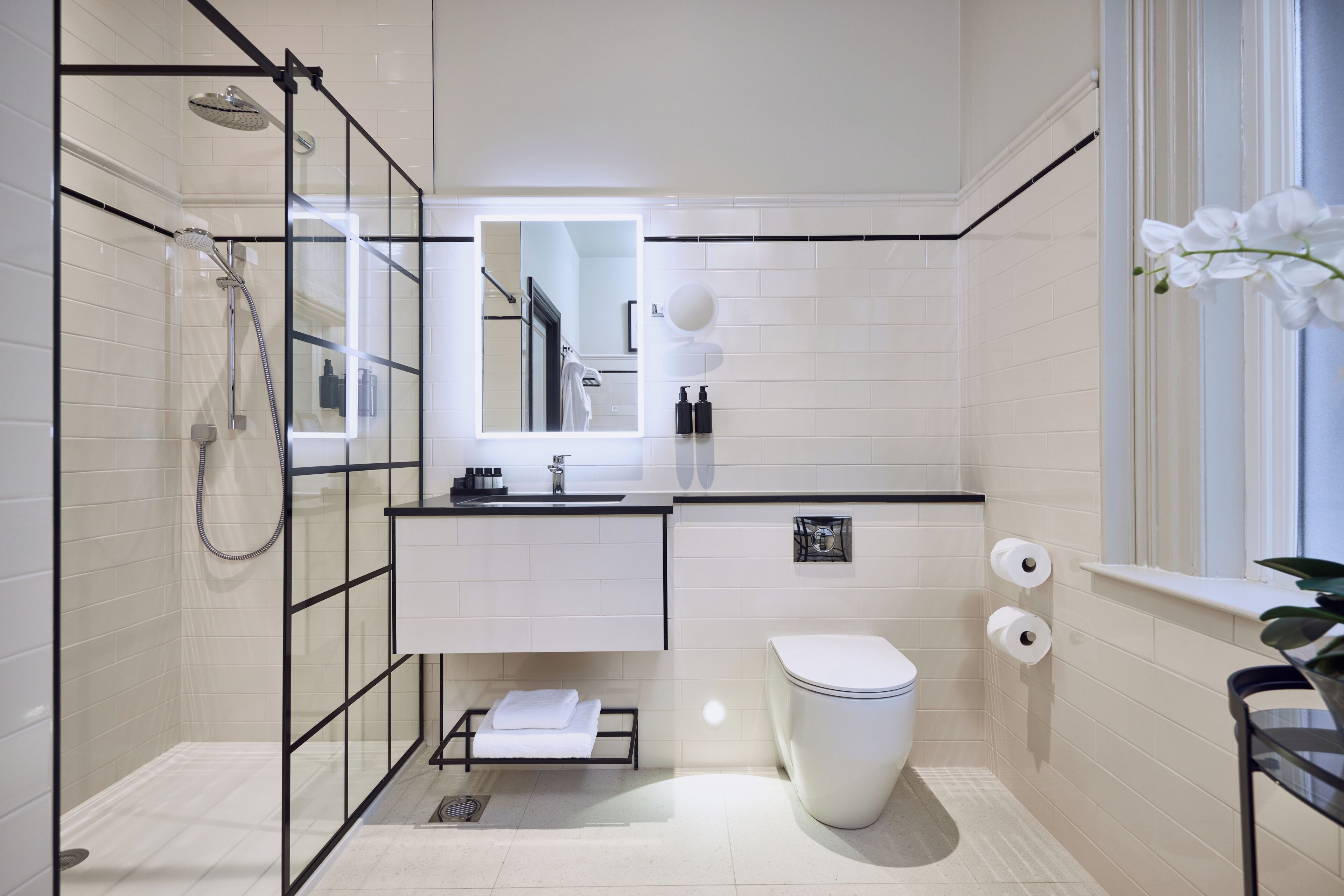 A modern bathroom with a glass shower enclosure, a toilet, a sink with a mirror, and a window with a plant.