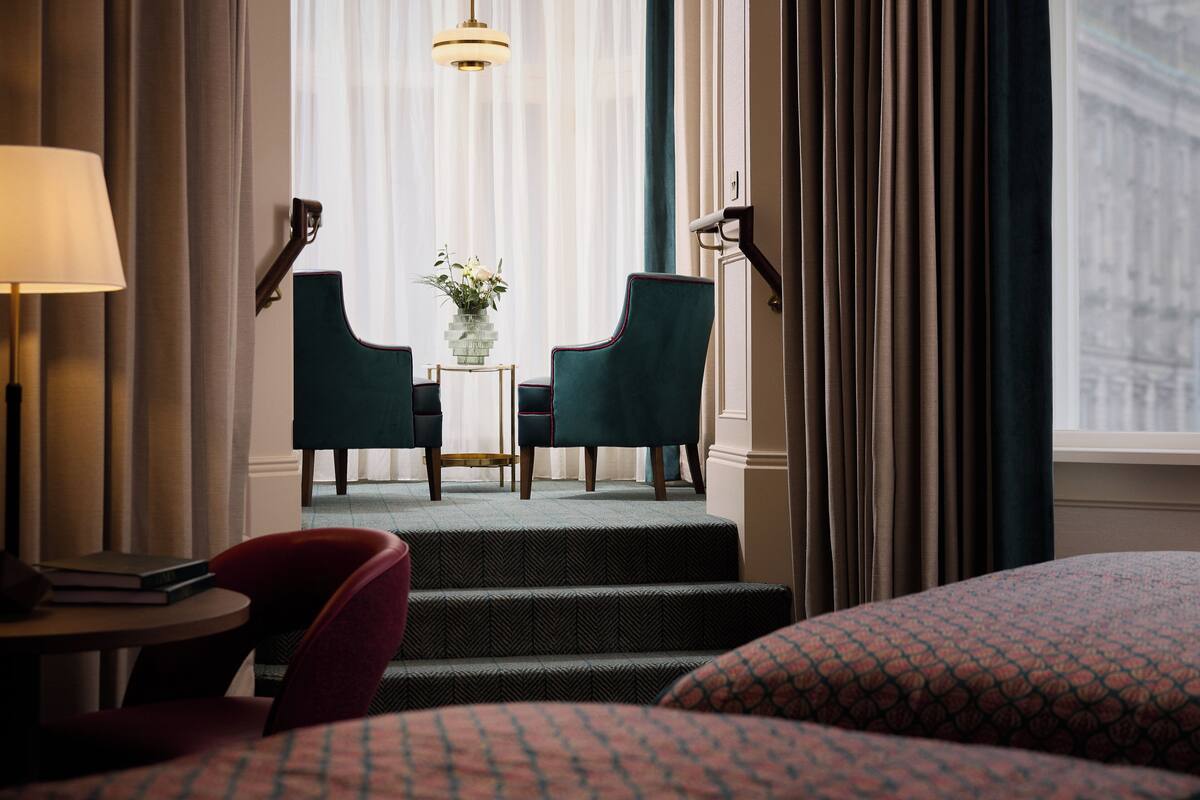 Une chambre d’hôtel avec un lit, un bureau avec une lampe, deux chaises, une petite table avec un vase et une vue sur un immeuble par la fenêtre.