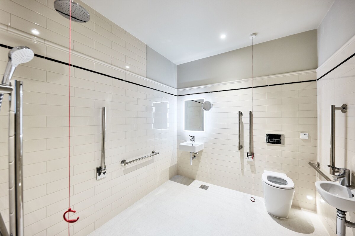 Un baño moderno con paredes de azulejos blancos, un espejo grande, un inodoro, un lavabo y una zona de ducha.