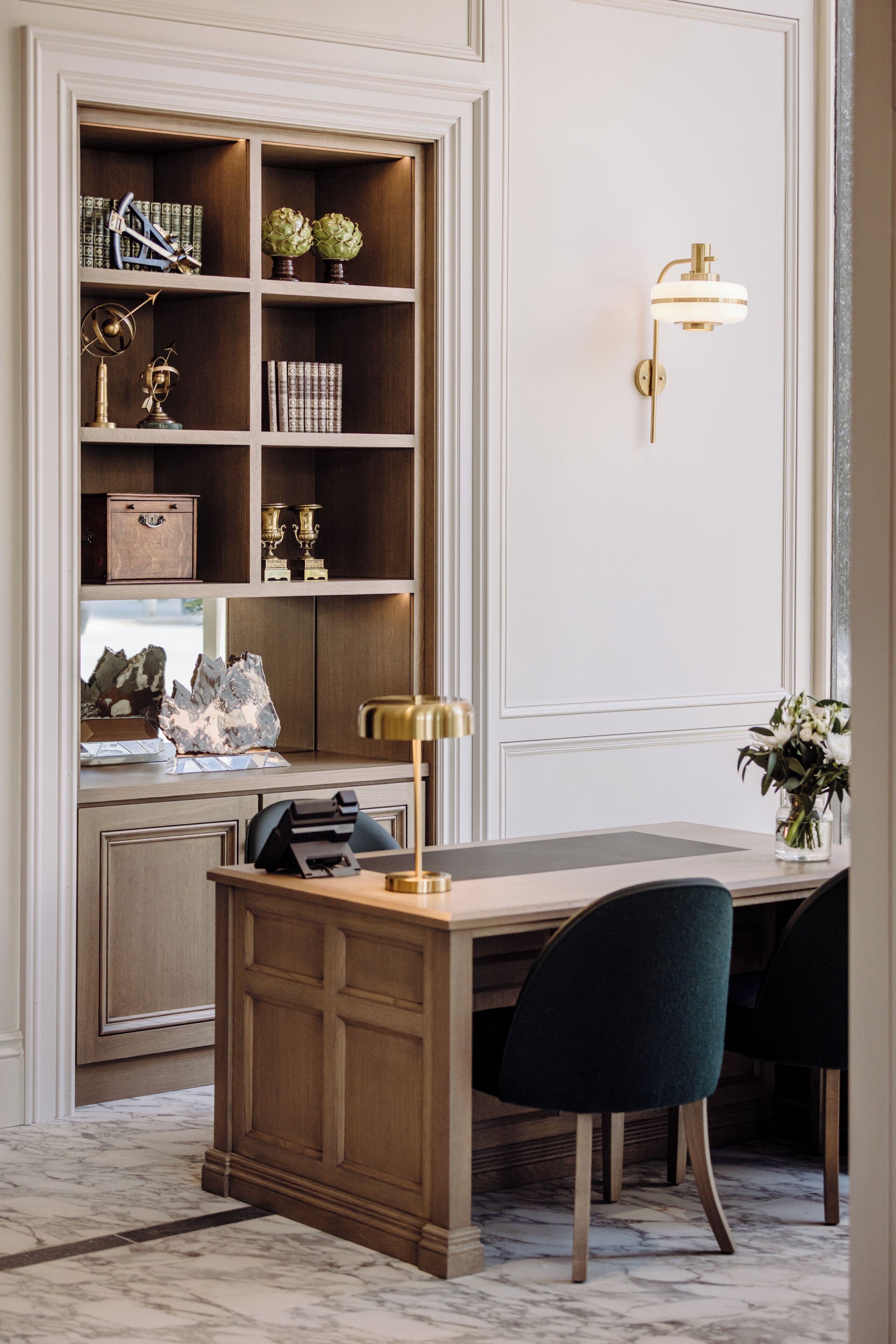 A study with a desk, two chairs, a bookshelf, and a wall-mounted lamp.