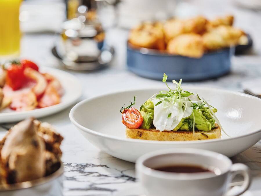A breakfast setting with toast topped with avocado and tomato, a cup of coffee, and a bowl of croissants.