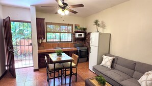 A spacious living area with a gray sofa, a dining table with chairs, a kitchen area with a microwave and refrigerator, and a ceiling fan. - Tiny Paradise Ground Floor Studio (Playas Del Coco)