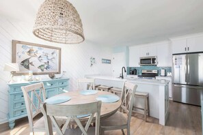 A modern kitchen with white cabinetry, a central island, stainless steel appliances, and a dining area with a wooden table and chairs. - New Listing! Water Views, Sunsets and Much More! (Kill Devil Hills)