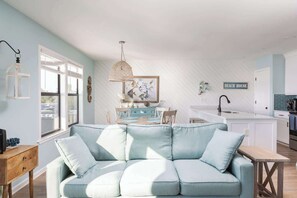 A modern kitchen with white cabinetry, a dining area with a light blue sofa, and a kitchen island. - New Listing! Water Views, Sunsets and Much More! (Kill Devil Hills)