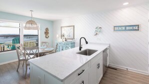 A kitchen with a large island, a dining area with chairs, a view of the water, and a 'Beach House' sign on the wall. - New Listing! Water Views, Sunsets and Much More! (Kill Devil Hills)