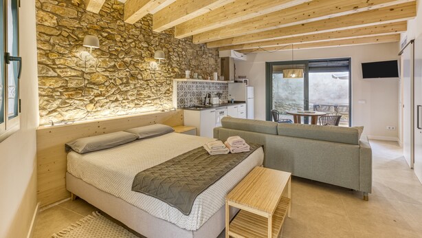 Een slaapkamer met een stenen muur, houten balken, een bed, een sofa, een klein tafeltje en een keukenhoek met een raam.