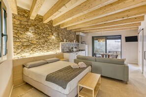 A bedroom with a stone wall, wooden beams, a bed, a sofa, a small table, and a kitchen area with a window. - Apartment 'Apartament Empordà' with Private Terrace, Wi-Fi and Air Conditioning (Peratallada)