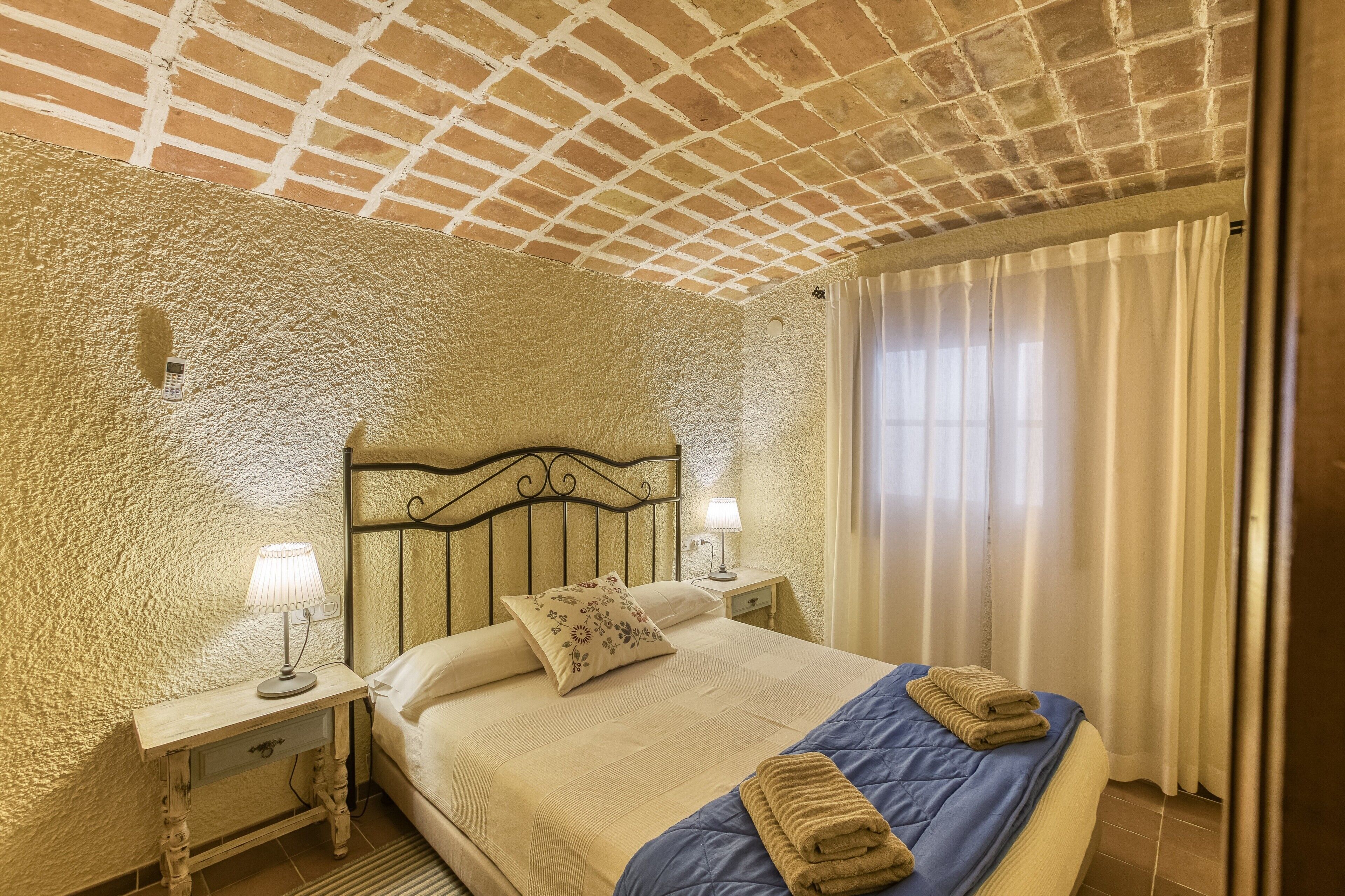 Una camera da letto con parete testurizzata, soffitto a motivi a mattoni, un letto con copriletto blu, due lampade da comodino e un comodino con una lampada.