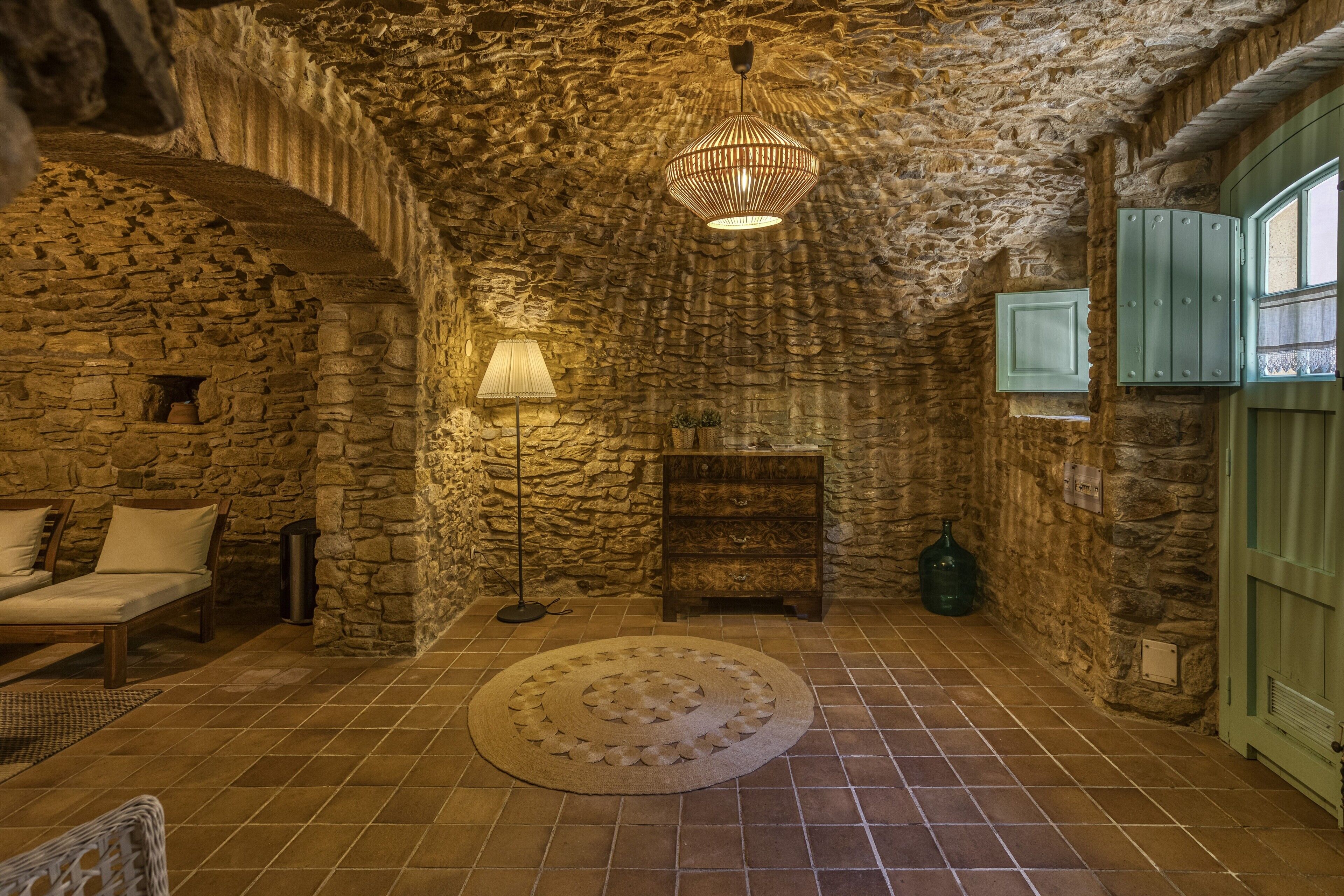 A stone-walled room with a bed, a floor lamp, a wooden cabinet, and a green door.