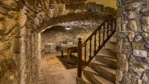 Un corridoio con pareti in pietra che conduce a una scala in legno, sullo sfondo si intravede un tavolo e delle sedie.