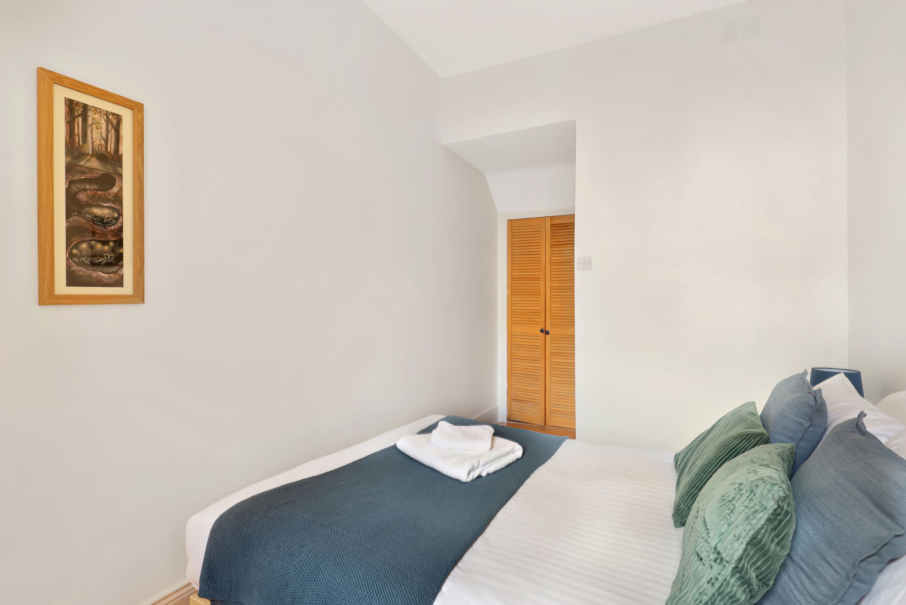 A neatly made bed with a blue blanket, a white towel, and multiple pillows. There is a framed picture on the wall and a wooden door in the background.