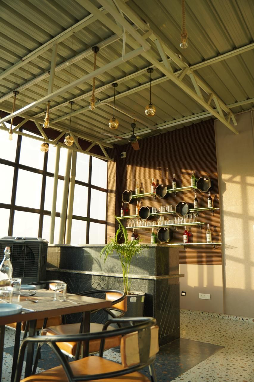 A restaurant with a high ceiling, hanging pendant lights, and a wall-mounted wine rack. Tables and chairs are arranged for dining.