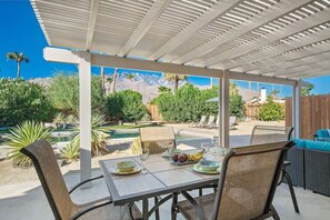 An outdoor patio with a table set for two, surrounded by chairs, a pool, and a mountain view.