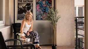 Une personne assise à une table, devant un livre et une tasse, avec une plante en pot et des affiches de bandes dessinées accrochées au mur.