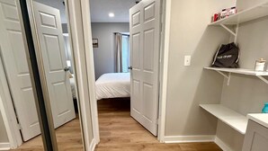 Ein Flur mit Holzboden, weißen Wänden und einem Bett im Hintergrund. Auf jeder Seite befinden sich weiße Türen und rechts eine Ablage mit einem Haartrockner.
