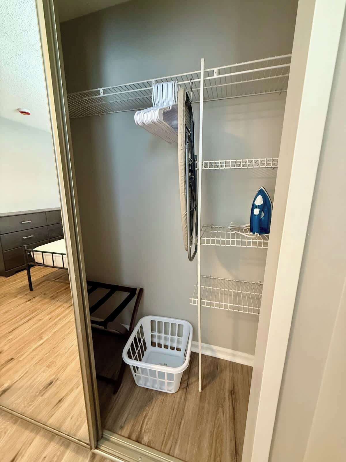 Habitación compacta con estructura de cama de metal, un cesto blanco para ropa y una estantería metálica con un objeto azul encima.