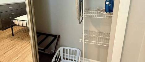 Habitación compacta con estructura de cama de metal, un cesto blanco para ropa y una estantería metálica con un objeto azul encima.