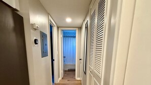 Un pasillo con piso de madera, paredes blancas y una puerta al final que da a una habitación con ventana y cortinas.