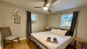 Un dormitorio con una cama, una silla, un ventilador de techo y dos ventanas con cortinas.