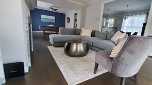 Ein modernes Wohnzimmer mit einem grauen Ecksofa, einem runden Metall-Couchtisch und einem grauen Sessel. Im Hintergrund ist ein Holzesstisch mit Stühlen zu sehen.