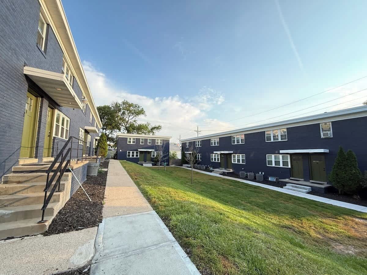 Uma fileira de edifícios de apartamentos modernos, com um caminho central, pequenas árvores e céu claro.