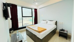 A hotel room with a bed, a desk, a chair, a nightstand, a TV, and a window with curtains. - Hotel Pleasure (Surat)