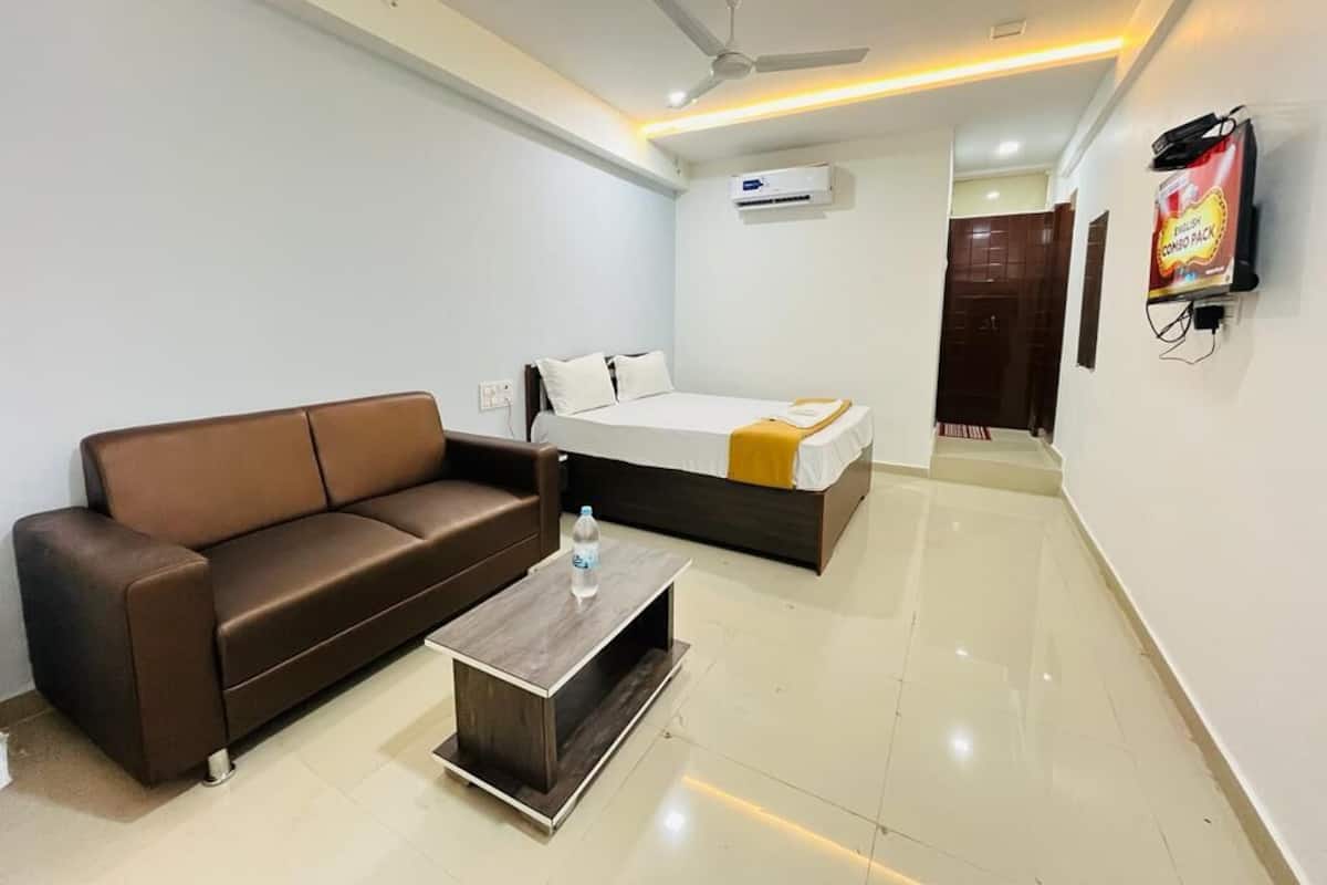 A modern hotel room with a brown sofa, a bed with a yellow throw, a wooden coffee table, an air conditioning unit, and a TV mounted on the wall.