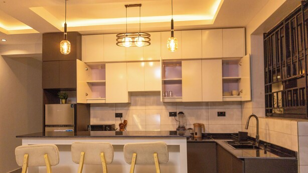 A modern kitchen with white cabinets, a central island with bar stools, pendant lighting, and a built-in stove. - Serene Apartment (Kigali)