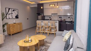 A modern living room with a sofa, two wooden coffee tables, and a kitchen area with bar stools. - Serene Apartment (Kigali)
